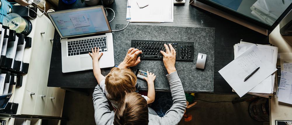 Ein erwachsener Mensch mit kurzen Haaren sitz an einem Schreibtisch und arbeitet am Computer. Auf dem Schoß hat er oder sie ein jüngeres Kind. Das Foto ist von oben aufgenommen, sodass man die Gesichter nicht sehen kann.