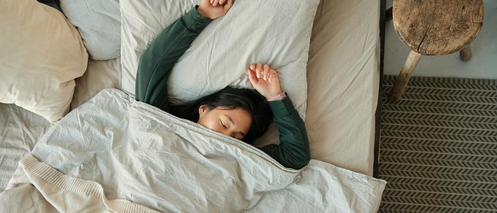 Eine junge Frau liegt rücklings mit ausgestreckten Armen auf dem Bett und schläft.