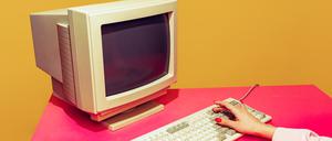 Ein Symbolbild zeigt einen alten Computer mit Tastatur, auf dem Hände etwas eingeben, auf einer rosafarbenen Tischdecke vor orangefarbenem Hintergrund.
