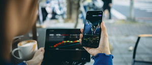 Eine Frau sitzt in einem Café und schaut auf Smartphone und Laptop Aktienkurse an.