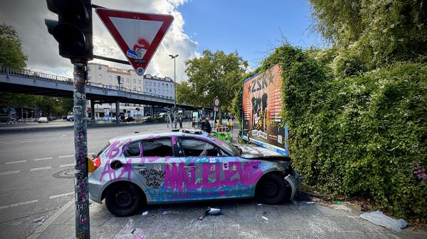Ein schwer beschädigter, silberner, mit Graffiti und Aufkleber versehener BMW 1 steht quer auf einem Gehweg neben einer Ampel in der Audre-Lorde-Straße Ecke Oranienstraße.