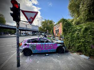 Ein schwer beschädigter, silberner, mit Graffiti und Aufkleber versehener BMW 1 steht quer auf einem Gehweg neben einer Ampel in der Audre-Lorde-Straße Ecke Oranienstraße.