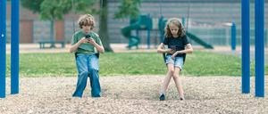 Zwei Kinder sitzen mit Smartphones in der Hand auf Schaukeln.