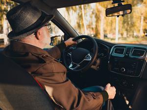 An elderly man wearing a hat enjoys a scenic urban drive during a sunny autumn day with vibrant foliage.