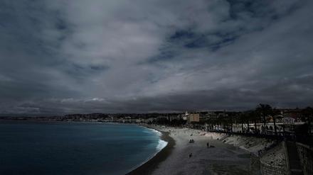 Menschen sitzen am Strand oder spazieren entlang der berühmten Promenade des Anglais unter einem bewölkten Himmel in Nizza. 
