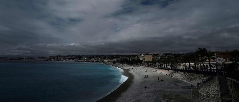 Menschen sitzen am Strand oder spazieren entlang der berühmten Promenade des Anglais unter einem bewölkten Himmel in Nizza. 