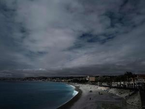 Menschen sitzen am Strand oder spazieren entlang der berühmten Promenade des Anglais unter einem bewölkten Himmel in Nizza. 