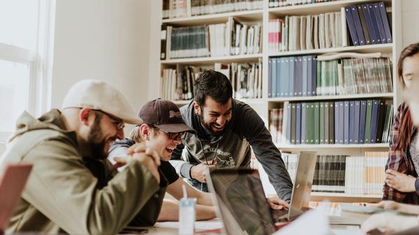 Drei junge Männer blicken in einem Büro gemeinsam in ein Laptop und freuen sich.