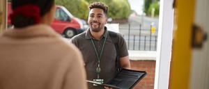 Ein lächelnder Firmenvertreter spricht mit der Bewohnerin eines Hauses an der Haustür. Um den Hals trägt er einen Ausweis an einem Band.