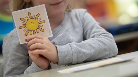 Illustration: Ein Mädchen hält in einer ersten Klasse an einer Grundschule mit Wechselunterricht eine Karte mit einer Sonne in der Hand. Die Karte hat sie ausgewählt, um zu zeigen, dass sie sich freut, wieder in der Schule zu sein. 
