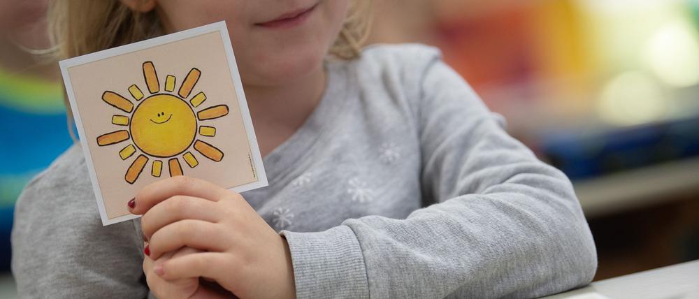 Illustration: Ein Mädchen hält in einer ersten Klasse an einer Grundschule mit Wechselunterricht eine Karte mit einer Sonne in der Hand. Die Karte hat sie ausgewählt, um zu zeigen, dass sie sich freut, wieder in der Schule zu sein.