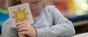 Illustration: Ein Mädchen hält in einer ersten Klasse an einer Grundschule mit Wechselunterricht eine Karte mit einer Sonne in der Hand. Die Karte hat sie ausgewählt, um zu zeigen, dass sie sich freut, wieder in der Schule zu sein. 