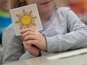 Illustration: Ein Mädchen hält in einer ersten Klasse an einer Grundschule mit Wechselunterricht eine Karte mit einer Sonne in der Hand. Die Karte hat sie ausgewählt, um zu zeigen, dass sie sich freut, wieder in der Schule zu sein. 