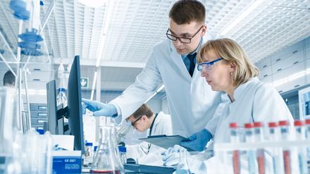 In einem modernen Labor führt eine erfahrene Wissenschaftlerin ein Gespräch mit einem jungen Laborassistenten. Er zeigt ihr Diagramme auf einem Klemmbrett, sie analysiert die Daten und gibt sie in ihren Computer ein.