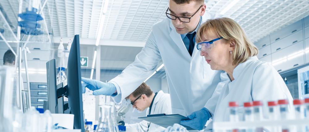 In einem modernen Labor führt eine erfahrene Wissenschaftlerin ein Gespräch mit einem jungen Laborassistenten. Er zeigt ihr Diagramme auf einem Klemmbrett, sie analysiert die Daten und gibt sie in ihren Computer ein.