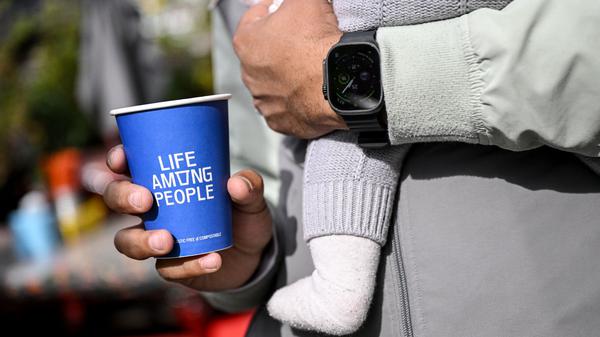 PRODUKTION - 25.09.2025, Berlin: Ein junger Mann hält einen Kaffeebecher der LAP-Kaffeekette in der Hand während er ein Baby auf dem Arm hat. (zu dpa: «Günstiger Kaffee im Trend - LAP Coffee fordert Szene heraus») Foto: Britta Pedersen/dpa +++ dpa-Bildfunk +++