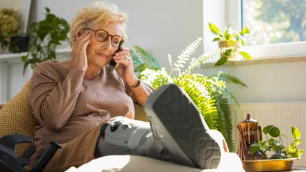 Eine ältere Frau sitzt telefonierend auf dem Sofa, während sie ihr Bein mit einer Unterschenkelorthese hochlagert.