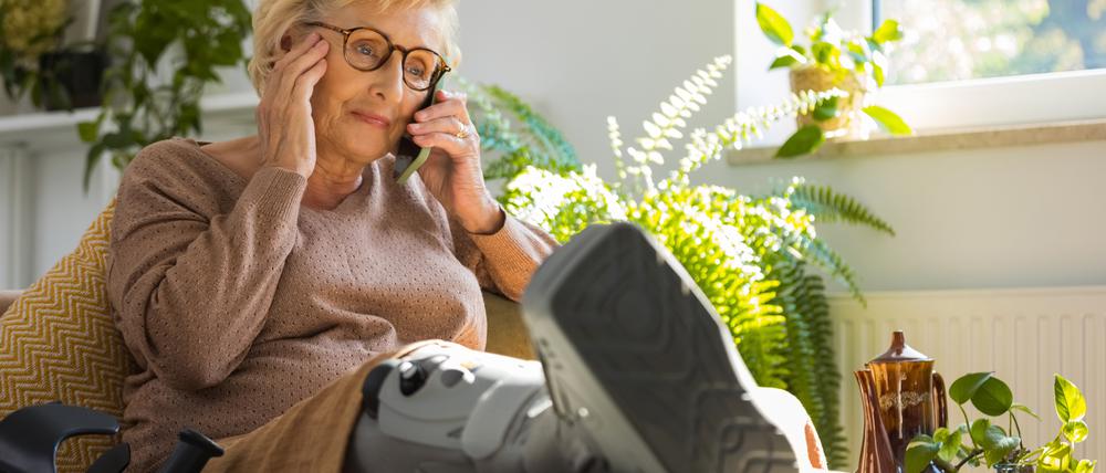 Eine ältere Frau sitzt telefonierend auf dem Sofa, während sie ihr Bein mit einer Unterschenkelorthese hochlagert.