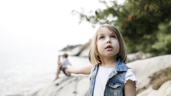 Ein etwa sechsjähriges Mädchen steht am Strand und schaut mit fragendem Blick in den Himmel.