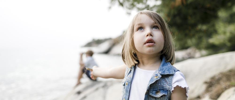 Ein etwa sechsjähriges Mädchen steht am Strand und schaut mit fragendem Blick in den Himmel.