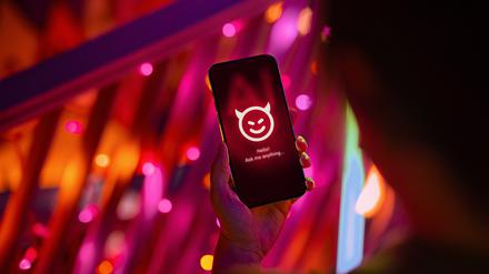 Eine Hand hält ein Smartphone mit einem Chatbot als lächelndes Teufelsgesicht.