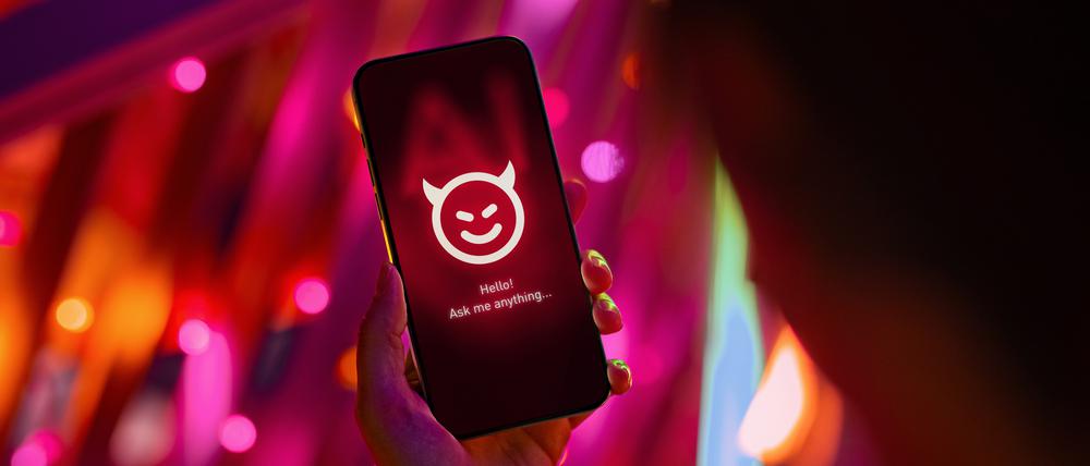 Eine Hand hält ein Smartphone mit einem Chatbot als lächelndes Teufelsgesicht.