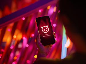 Eine Hand hält ein Smartphone mit einem Chatbot als lächelndes Teufelsgesicht.