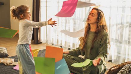 Eine Mutter sitzt erschöpft mit geschlossenen Augen auf dem Fußboden, während ein etwa fünfjähriges Mädchen sie mit buntem Bastelpapier bewirft.