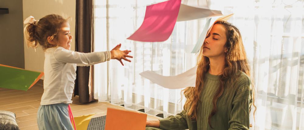 Eine Mutter sitzt erschöpft mit geschlossenen Augen auf dem Fußboden, während ein etwa fünfjähriges Mädchen sie mit buntem Bastelpapier bewirft.