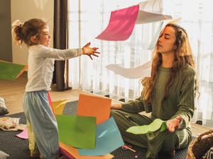 Eine Mutter sitzt erschöpft mit geschlossenen Augen auf dem Fußboden, während ein etwa fünfjähriges Mädchen sie mit buntem Bastelpapier bewirft.