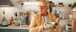 Ein älterer Mann isst im Stehen in seiner Küche ein Müsli.