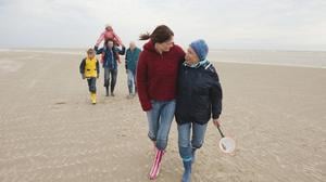 Eine junge und eine ältere Frau laufen Arm in Arm am Strand entlang.