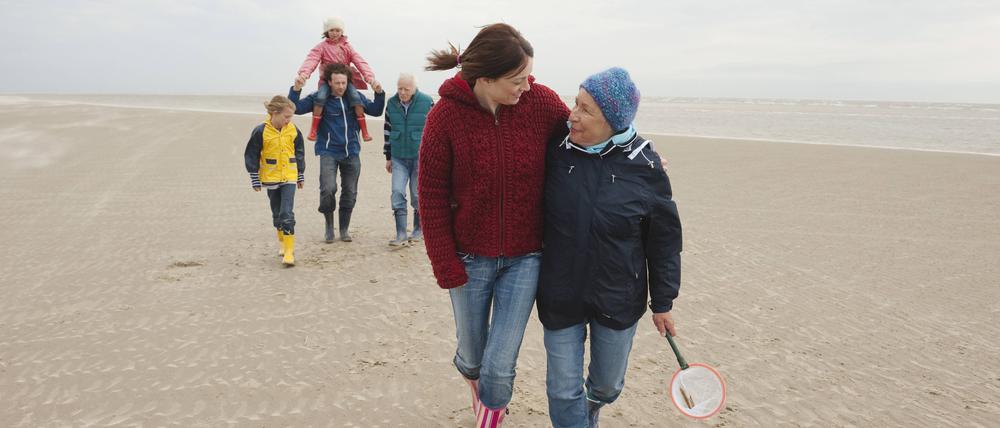Eine junge und eine ältere Frau laufen Arm in Arm am Strand entlang.