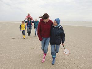 Eine junge und eine ältere Frau laufen Arm in Arm am Strand entlang.
