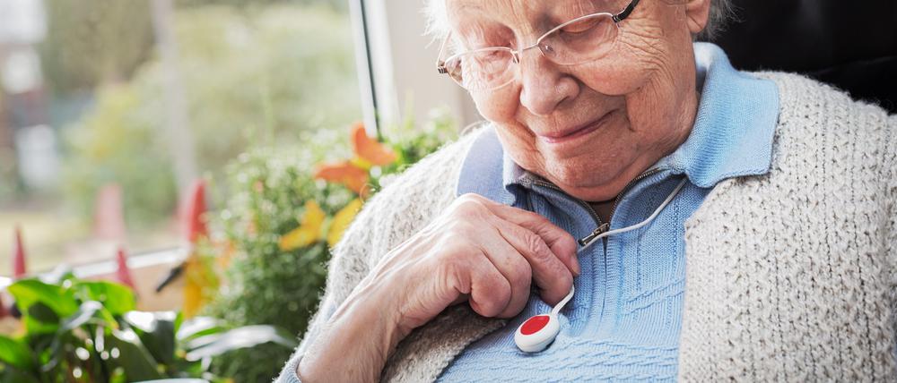 Eine ältere Dame blickt auf den Notrufknopf, den sie an einer Kette um den Hals trägt.