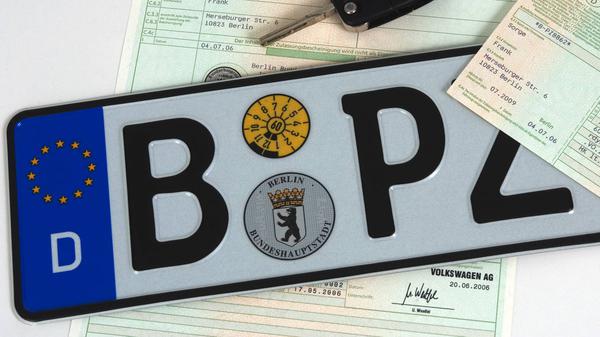 Ein Symbolbild zeigt ein Berliner Autokennzeichen, einen Autoschlüssel und einen Fahrzeugbrief. 