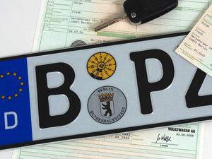 Ein Symbolbild zeigt ein Berliner Autokennzeichen, einen Autoschlüssel und einen Fahrzeugbrief.