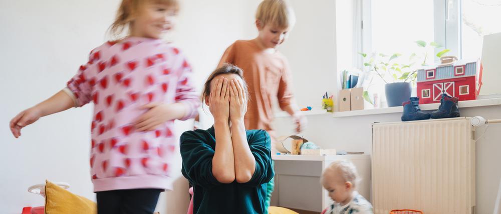 Eine gestresste Mutter hält die Hände vor ihr Gesicht, um sie herum spielen drei Kleinkinder.