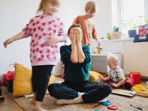 Eine gestresste Mutter hält die Hände vor ihr Gesicht, um sie herum spielen drei Kleinkinder.