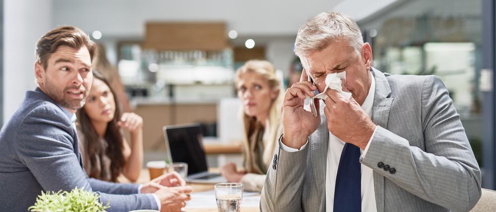 Ein Mann im Anzug niest in ein Taschentuch, während seine Kollegen im Hintergrund ihn erschrocken anschauen (Symbolfoto).