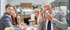 Ein Mann im Anzug niest in ein Taschentuch, während seine Kollegen im Hintergrund ihn erschrocken anschauen (Symbolfoto).