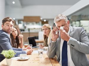 Ein Mann im Anzug niest in ein Taschentuch, während seine Kollegen im Hintergrund ihn erschrocken anschauen (Symbolfoto).