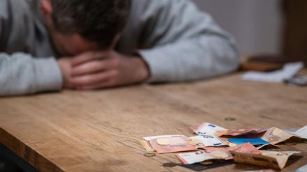 Ein Mann hat seinen Kopf auf seine Hände gelegt, während sich vor ihm Geldscheine, Münzen und Bankkarten auf einem Tisch befinden.