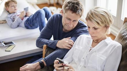 Eine Mutter und ein Vater blicken besorgt auf einen Handybildschirm. Im Hintergrund ist ein Kind zu sehen.
