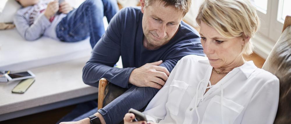 Eine Mutter und ein Vater blicken besorgt auf einen Handybildschirm. Im Hintergrund ist ein Kind zu sehen.