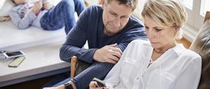 Eine Mutter und ein Vater blicken besorgt auf einen Handybildschirm. Im Hintergrund ist ein Kind zu sehen.