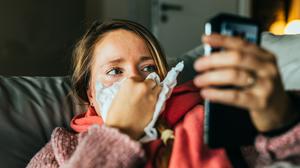 Eine Frau liegt erkältet auf dem Sofa und hält ihr Smartphone in der Hand.