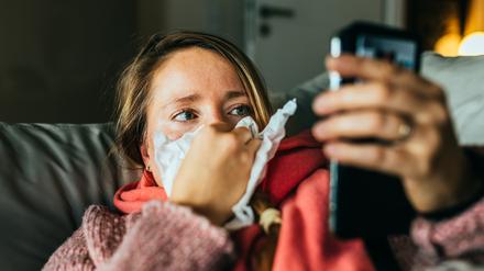 Eine Frau liegt erkältet auf dem Sofa und hält ihr Smartphone in der Hand.