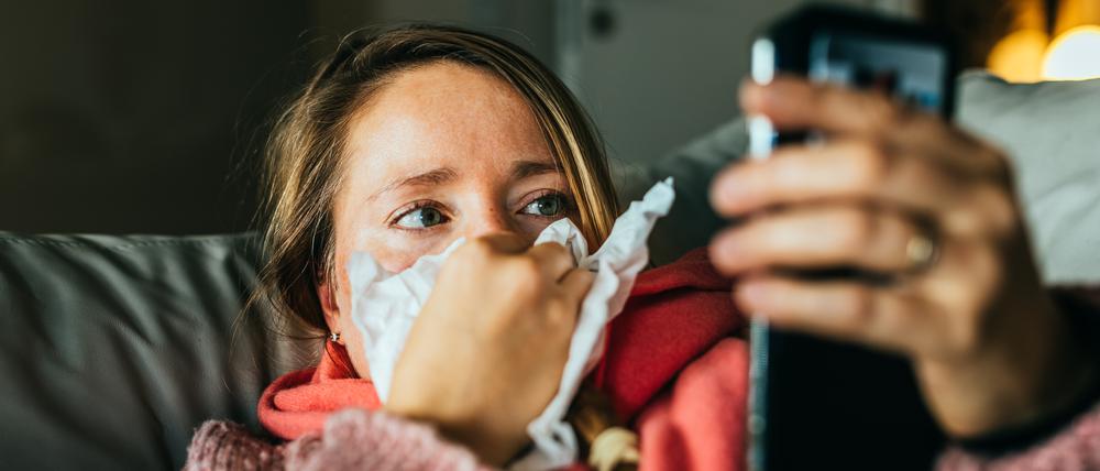 Eine Frau liegt erkältet auf dem Sofa und hält ihr Smartphone in der Hand.