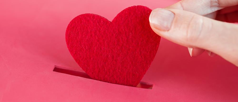 Ein rotes Herz wird in eine Spendenbox gesteckt.